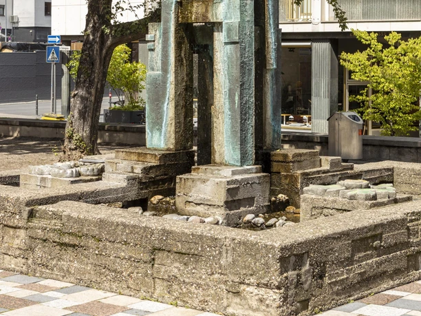 Theo-Burauen-Brunnen Das Bild zeigt den Theo-Burauen-Brunnen, einen eleganten, vertikalen Steingussbrunnen in Köln. Umliegend sind ein grüner Baum und moderne Gebäude sichtbar, die das urbane Stadtbild ergänzen.