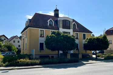 Gelbes zweistöckiges Gebäude mit rotem Dach und Turmuhr, umgeben von Bäumen und Sträuchern, bei sonnigem Wetter.