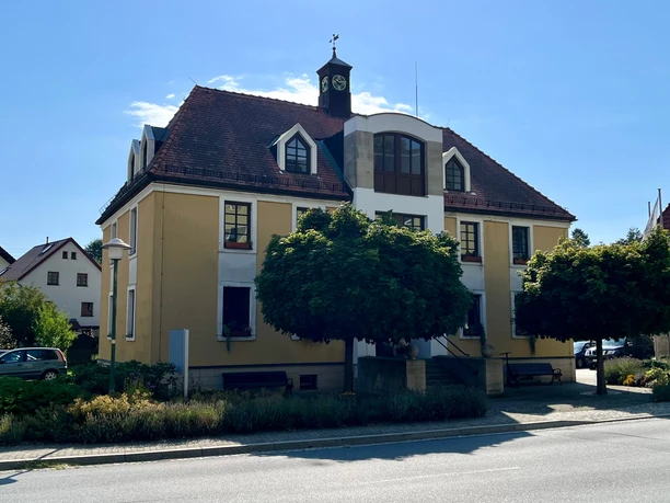 Gelbes zweistöckiges Gebäude mit rotem Dach und Turmuhr, umgeben von Bäumen und Sträuchern, bei sonnigem Wetter.