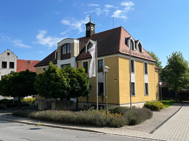 Gelbes Gebäude mit rotem Ziegeldach und Uhrturm, umgeben von Bäumen und Sträuchern unter blauem Himmel.