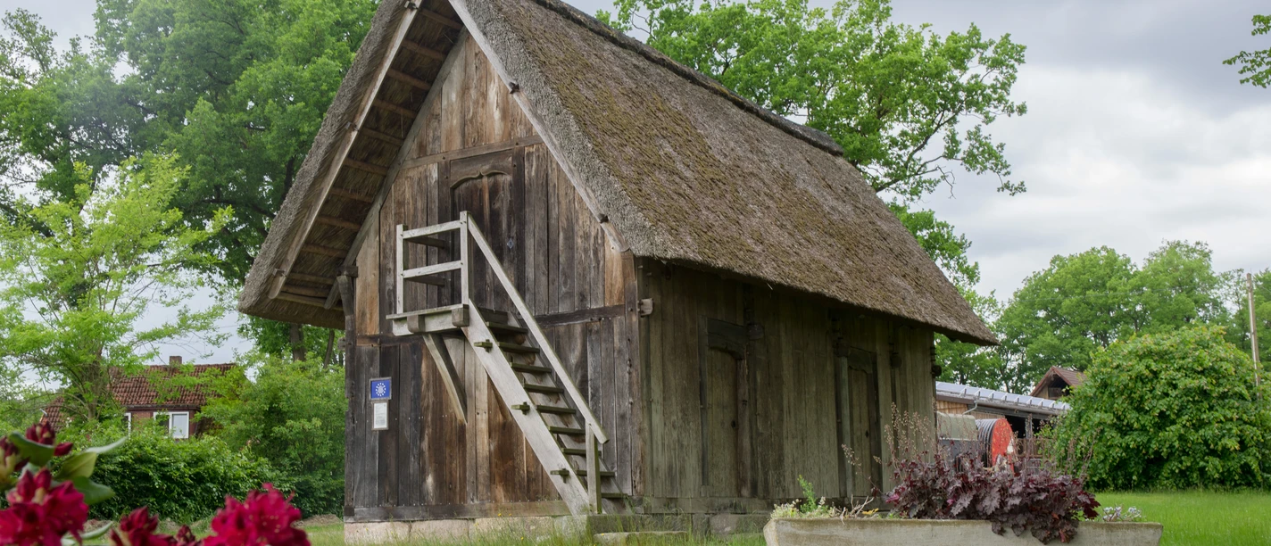 speicher-volkwardingen Historischer Treppenspeicher mit Reetdach auf grüner Wiese, umgeben von Bäumen und Blumen.