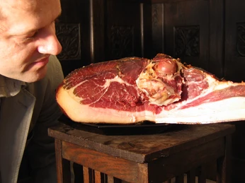 Schinkenanschnitt Ein Mann betrachtet aufmerksam einen frisch angeschnittenen großen Schinken. Der Schinken ruht auf einem Holztisch und zeigt seine saftige, rote Innenfläche mit einer dünnen Fettschicht am Rand. Der Mann trägt ein weißes Hemd und steht nah am Schinken, was sein Interesse am Produkt deutlich macht.
