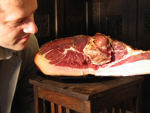 Schinkenanschnitt Ein Mann betrachtet aufmerksam einen frisch angeschnittenen großen Schinken. Der Schinken ruht auf einem Holztisch und zeigt seine saftige, rote Innenfläche mit einer dünnen Fettschicht am Rand. Der Mann trägt ein weißes Hemd und steht nah am Schinken, was sein Interesse am Produkt deutlich macht.