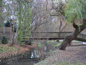 Herbstliche Szene in einem Park mit einer hölzernen Brücke über einem kleinen Bach, umgeben von Bäumen.