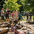 Kinder spielen auf einem Wasserspielplatz umgeben von Bäumen, während Eltern zusehen.