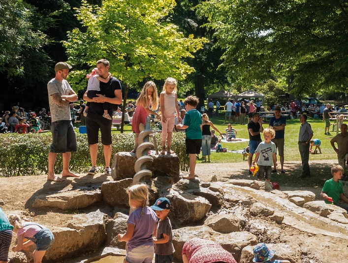Kinder spielen auf einem Wasserspielplatz umgeben von Bäumen, während Eltern zusehen.