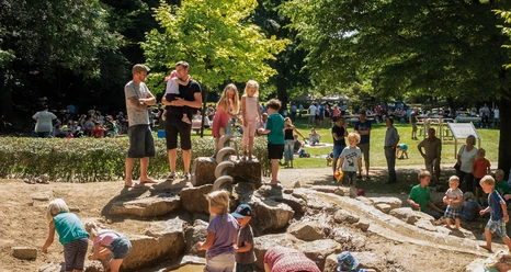 Wasserspielplatz Maiwiese Kinder spielen auf einem Wasserspielplatz umgeben von Bäumen, während Eltern zusehen.