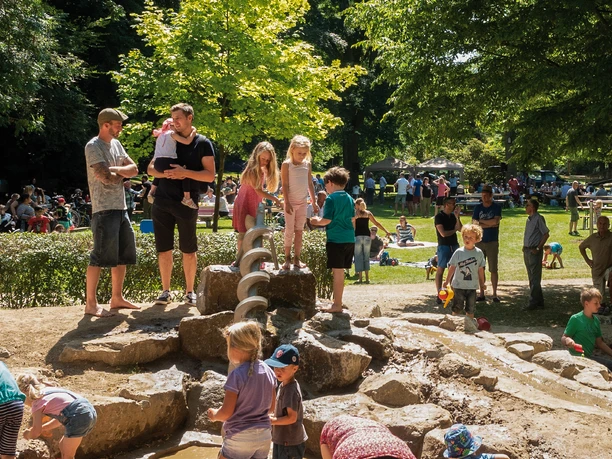 Wasserspielplatz Maiwiese Kinder spielen auf einem Wasserspielplatz umgeben von Bäumen, während Eltern zusehen.