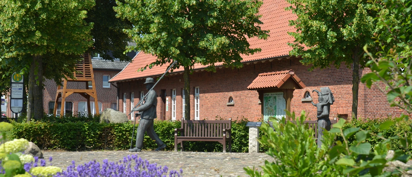 Krulls Hus Backsteingebäude mit rotem Dach, flankiert von Bäumen, Blumenbeet und Bronzefiguren im Vordergrund.