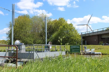 Kurbelfähre Holtland Die Kurbelfähre Holtland überquert einen Kanal, umgeben von grüner Vegetation. Ein modernes Windrad ist im Hintergrund.