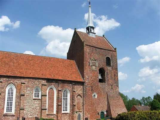Ev.-ref. Kirche Groothusen Backsteinkirche mit hohem Turm und spitzem Dachhelm, roten Ziegeln und gotischen Fenstern unter blauem Himmel.