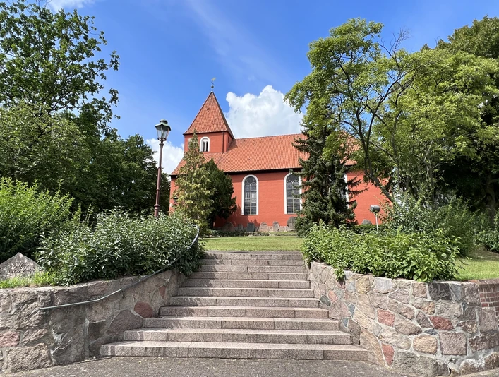 Die St. Petri-Kirche in Kirchlinteln mit roter Fassade, umgeben von Bäumen und eingebettetem Treppenaufgang.