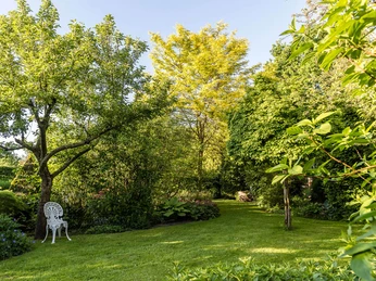 garten-landhof-tausendschoen Ein idyllischer Garten mit saftigem Grün, bunten Blumen und einem weißen Gartenstuhl.