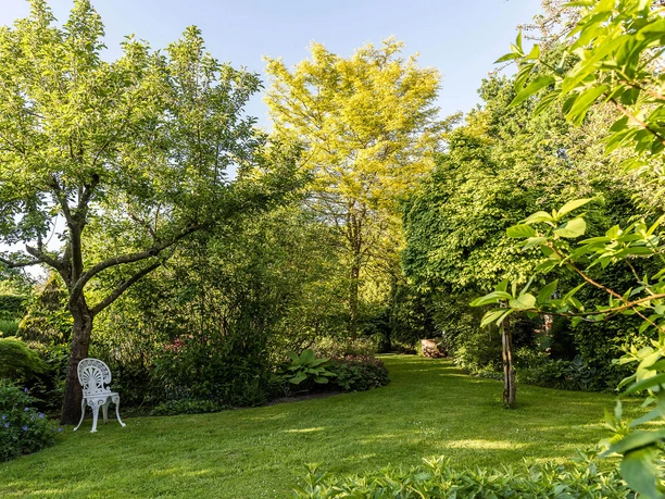 garten-landhof-tausendschoen Ein idyllischer Garten mit saftigem Grün, bunten Blumen und einem weißen Gartenstuhl.