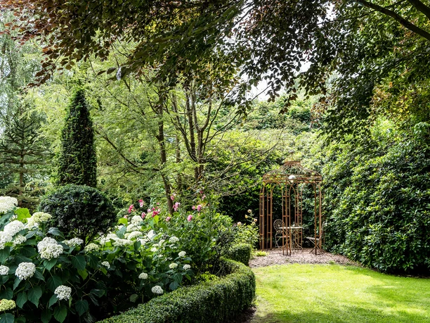 garten-landhof-tausendschoen-apen-5 Ein liebevoll gepflegter Garten mit farbenfrohen Blumenbeeten, einem grünen Rasen und einer eleganten Metalllaube, die von Rankgewächsen umgeben ist. Üppig blühende Hortensien im Vordergrund, dichter Baum- und Sträucherbestand im Hintergrund.