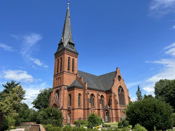 Backsteinkirche St. Jakobi in Wittlohe mit spitzem Turm vor blauem Himmel, umgeben von üppigem Grün.