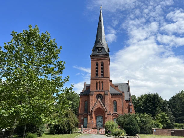 A brick church with a tall, pointed tower, nestled in the countryside under a blue sky in Wittlohe.