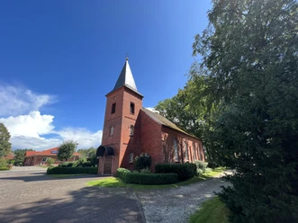 Backsteinkirche mit hohem Turm, umgeben von grünen Bäumen und einem gepflasterten Vorplatz.