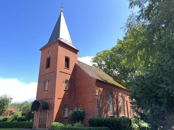 Rotgeklinkerte Kirche mit spitzem Turmdach und schlichten Rundbogenfenstern, umgeben von Bäumen.