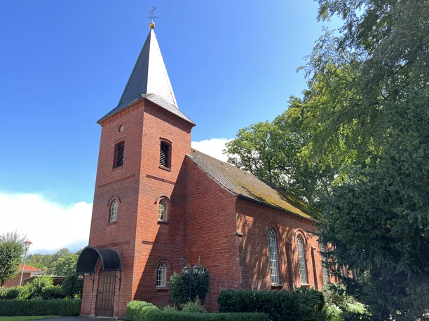 Rotgeklinkerte Kirche mit spitzem Turmdach und schlichten Rundbogenfenstern, umgeben von Bäumen.