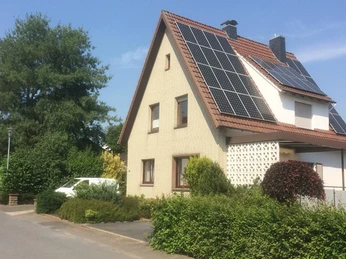 Ein zweistöckiges Wohnhaus mit Solarpaneelen auf dem Dach, umgeben von grünen Sträuchern.