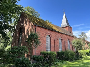 St. Matthäus-Kirche Brunsbrock Backsteinkirche mit Satteldach und Turmspitze, umgeben von Bäumen und gepflegtem Grün unter blauem Himmel.