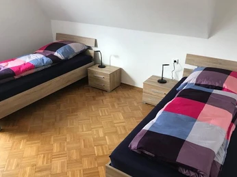 Zwei Einzelbetten mit bunter Bettwäsche, Holzboden und Nachttischen mit Lampen, Fenster nicht sichtbar.