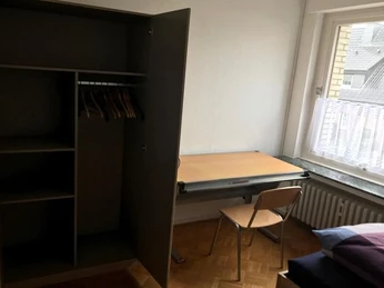 Ein schlichtes Zimmer mit Kleiderschrank, Schreibtisch und Stuhl, daneben ein Fenster mit Tageslicht.