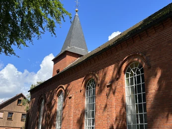 Backsteinbau mit Turm und Spitzdach, große Fenster mit Rundbögen, blauer Himmel, umgeben von Bäumen.