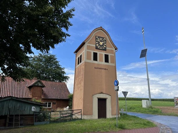 Glockenturm in Neddenaverbergen Glockenturm in Neddenaverbergen, Rotklinkerbau mit Uhr, umgeben von ländlicher Umgebung und blauem Himmel.