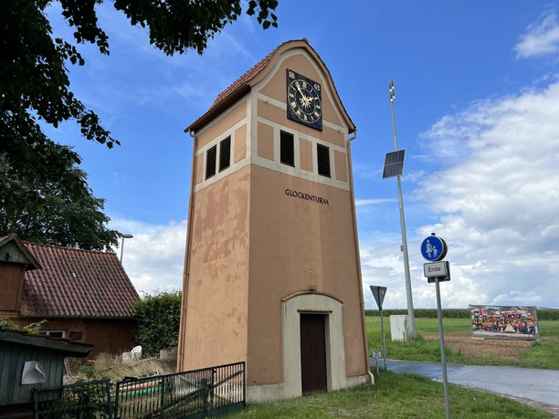 A historic bell tower with a red roof and a clock, surrounded by rural scenery and signposts.