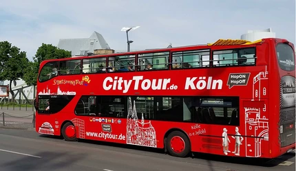 Cologne CityTour A red double-decker "CityTour Köln" bus is parked on the side of the road with its upper deck open. The bus is adorned with white illustrations of Cologne landmarks.