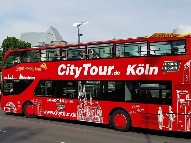 Cologne CityTour A red double-decker "CityTour Köln" bus is parked on the side of the road with its upper deck open. The bus is adorned with white illustrations of Cologne landmarks.