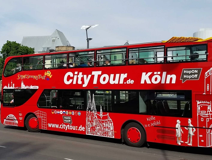 Cologne CityTour Ein roter Doppeldecker-Bus der „CityTour Köln“ steht am Straßenrand, mit geöffnetem Oberdeck. Die weißen Illustrationen von Kölner Wahrzeichen zieren den Bus.A red double-decker "CityTour Köln" bus is parked on the side of the road with its upper deck open. The bus is adorned with white illustrations of Cologne landmarks.