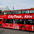 Kölner CityTour Ein roter Doppeldecker-Bus der „CityTour Köln“ steht am Straßenrand, mit geöffnetem Oberdeck. Die weißen Illustrationen von Kölner Wahrzeichen zieren den Bus.A red double-decker "CityTour Köln" bus is parked on the side of the road with its upper deck open. The bus is adorned with white illustrations of Cologne landmarks.