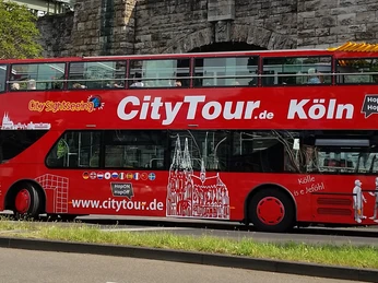 Cologne CityTour Ein knallroter Doppeldeckerbus der Kölner CityTour fährt vor einer Steinmauer entlang. Der Bus ist mit weißen Grafiken und dem Schriftzug "CityTour.de Köln" verziert.A bright red double-decker bus from Cologne CityTour drives along in front of a stone wall. The bus is decorated with white graphics and the lettering "CityTour.de Köln".