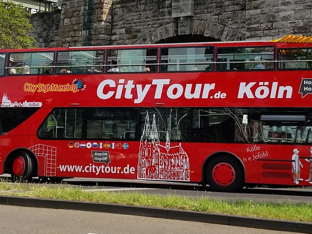 Kölner CityTour Ein knallroter Doppeldeckerbus der Kölner CityTour fährt vor einer Steinmauer entlang. Der Bus ist mit weißen Grafiken und dem Schriftzug "CityTour.de Köln" verziert.
