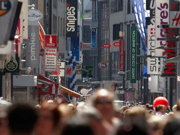 Hohen Straße Hohe Straße in Köln, belebt mit Einkaufenden. Verschiedene Geschäfte und farbige Werbeschilder flankieren den belebten Weg. Direkte Straßenansicht mit im Hintergrund sichtbaren Gebäuden.