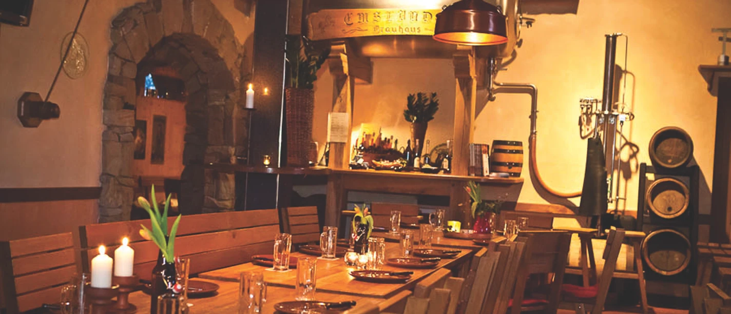 Landhaus Brauerei Borchert Holzverkleideter Gastraum mit langen Tischen, Kupferlampen, gedecktem Tisch und Brauereiausrüstung.