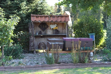 fehn-imkerei-lind-03-emsland-tourismus-christina-poll Holzüberdachung mit Bienenkörben, bunten Beuten und Dekofigur einer Biene im grünen Gartenbereich