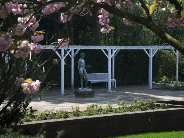 Freilichtbühne Bronzestatue eines stehenden Mannes hinter laubbedecktem Baum, umrahmt von Rosablüten und Pergola.