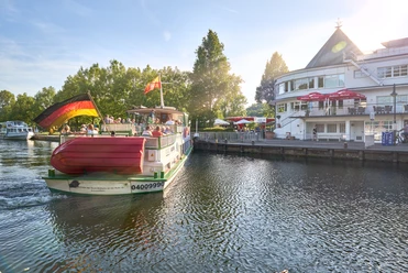 Wasserbahnhof, Mülheim an der Ruhr Ein Passagierschiff mit Deutschlandflagge an einem Flussanleger, flankiert von Bäumen und einem Gebäude.