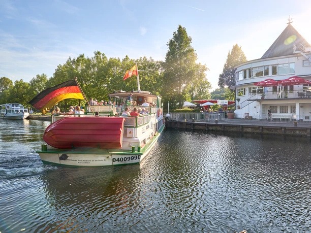 Wasserbahnhof, Mülheim an der Ruhr Ein Passagierschiff mit Deutschlandflagge an einem Flussanleger, flankiert von Bäumen und einem Gebäude.