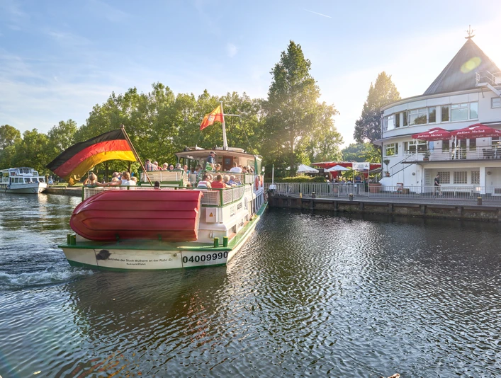 Wasserbahnhof, Mülheim an der Ruhr Ein Passagierschiff mit Deutschlandflagge an einem Flussanleger, flankiert von Bäumen und einem Gebäude.