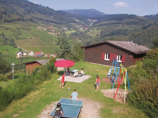 Im Hintergrund dunkelbraunes Holzhaus mit roten Fensterläden, davor Sitzplatz, Schaukel, Kind an Tischtennisplatte