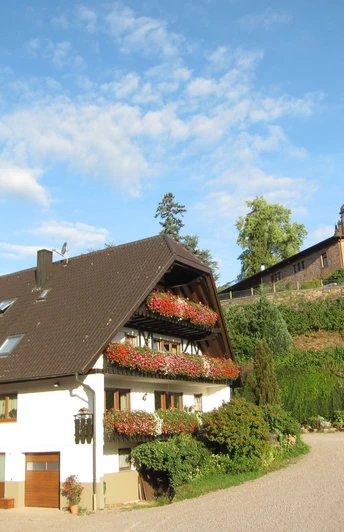 Ein vierstöckiges weißes Haus mit braunem Dach, an den Balkonen rote Blumen. Im Hintergrund ein altes Schloss