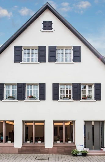 Hausansicht. Eine dreistöckige, weiße Hausfassade. Im Erdgeschoss drei große Fenster, darüber Fenster mit Fensterläden