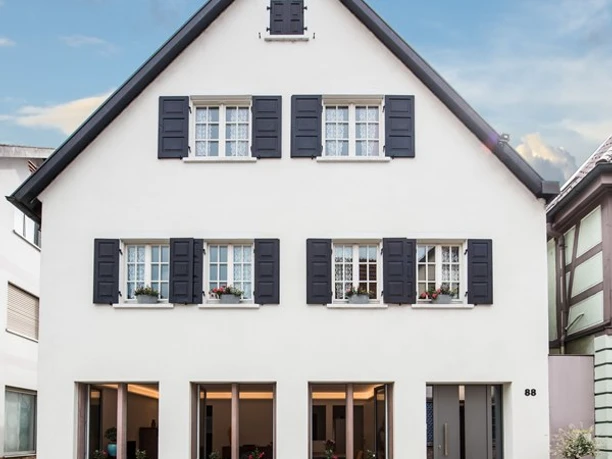 Hausansicht. Eine dreistöckige, weiße Hausfassade. Im Erdgeschoss drei große Fenster, darüber Fenster mit Fensterläden