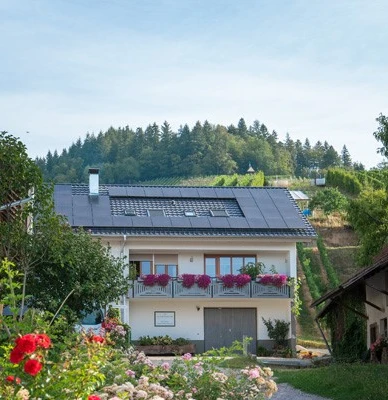 Dreistöckiges weißes Haus mit grauem Dach, Balkon mit Blumen. Davor ein Hof mit Sträuchern
