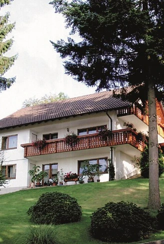 Ein zweistöckiges weißes Haus mit braunem Dach und großem Holzbalkon. Im Vordergrund eine Rasenfläche mit Nadelbäumen